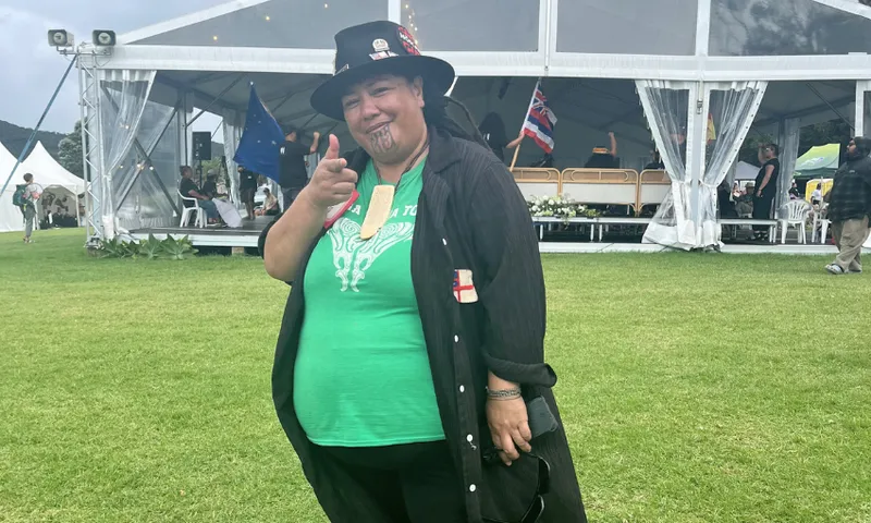 A person wearing a black hat, black shirt, and green t-shirt stands on grass, smiling and pointing at the camera. Behind them is a large white event tent with people and flags visible inside.