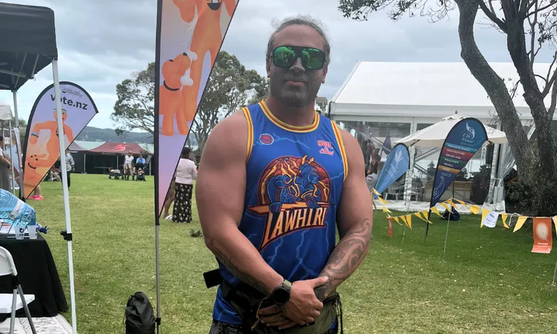 A man with sunglasses and a ponytail stands outdoors at an event, wearing a blue basketball jersey that says “TAWHIRI.” Behind him are tents, banners, trees, and people on a grassy area.