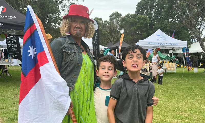 A woman with a traditional moko and red hat holds a Samoan flag, standing next to two smiling boys at an outdoor cultural festival with tents and trees in the background.