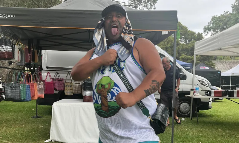 A man in a white basketball jersey poses playfully, sticking out his tongue and holding a bag over his shoulder at an outdoor market with tents and colorful bags in the background.