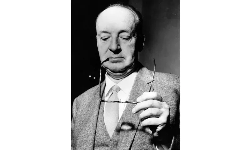 A black and white photograph of the writer Vladimir Nabokov who is holding out his glasses and looking down at them. He has a bald head and is wearing a suit.