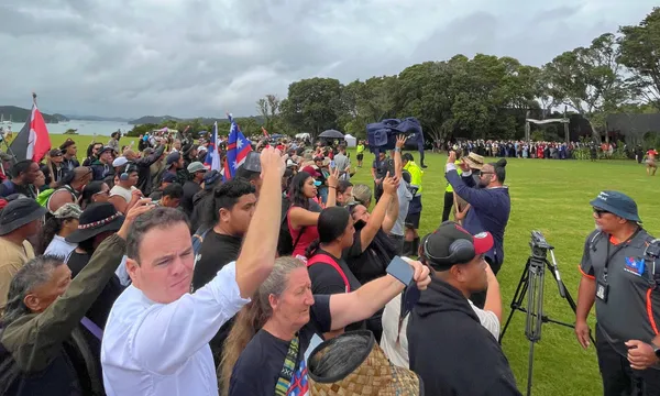 A large crowd stands outdoors on a grassy field, many raising their fists or holding phones. Some people wave flags. Trees and a cloudy sky are in the background. A few security personnel and a camera are visible on the right.
