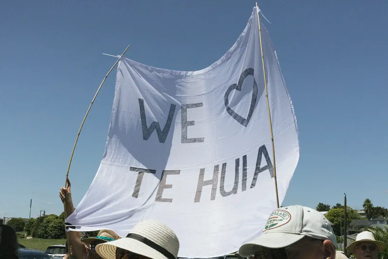 A group of people outdoors hold up a large white banner with the words "WE ♥ TE HUIA" written in bold black letters on a sunny day. The sky is clear and blue above them.