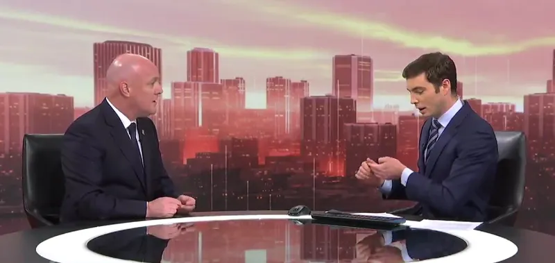 Two men in suits sit across from each other at a news desk with a city skyline backdrop. One speaks while the other looks down at his notes. A mouse and tablet are on the desk. The lighting is warm and professional.