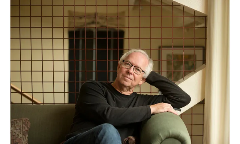 A photo of the poet Bill Manhire who is sitting on a green couch, in a relaxed pose. He is wearing a dark jumper and is slightly smiling.