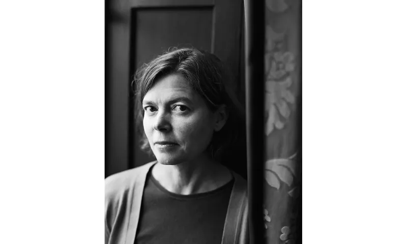 A black and white photo of the poet Jenny Bornholdt. She is leaning against a wall and looking into the camera with her face slightly facing away. Her hair is in a short bob, she wears a top and cardigan.