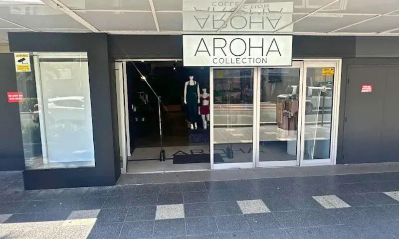 Storefront of Aroha Collection with glass doors, mannequins displaying clothing inside, and a large white sign above the entrance. The sidewalk and street reflections are visible in the glass.