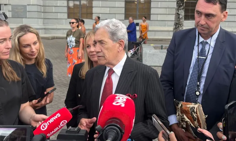 Winston Peters looks to his right while standing in front of a row of microphones, next to MPs Jenny Marcroft and Jamie Arbuckle.