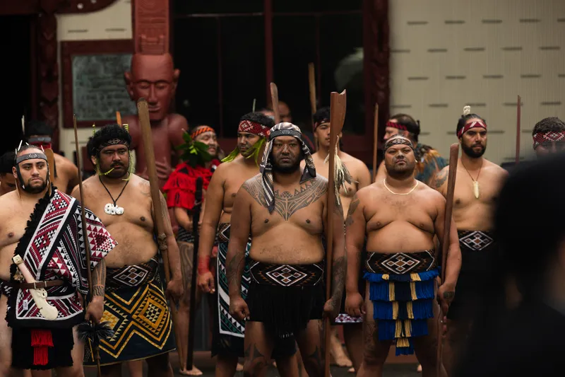 A group of Māori men in traditional clothing and accessories stand in formation outdoors, some holding wooden weapons, with a carved wooden figure and building in the background.
