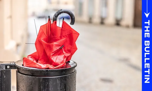 A crumpled red umbrella is discarded inside a black trash can on a city street. Buildings and pavement are out of focus in the background. A blue vertical banner on the right reads "THE BULLETIN.