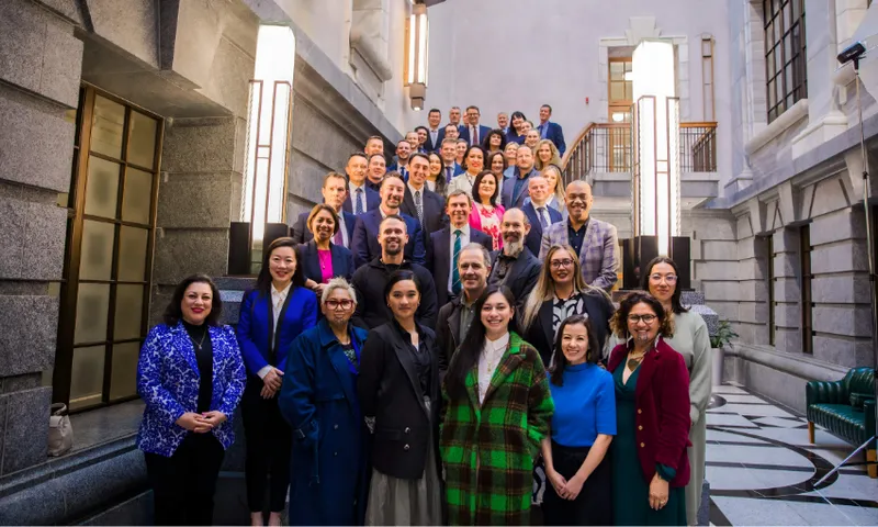 A large group of professionally dressed people stand on a grand staircase in a bright, elegant building, smiling at the camera. The group includes men and women of diverse backgrounds.