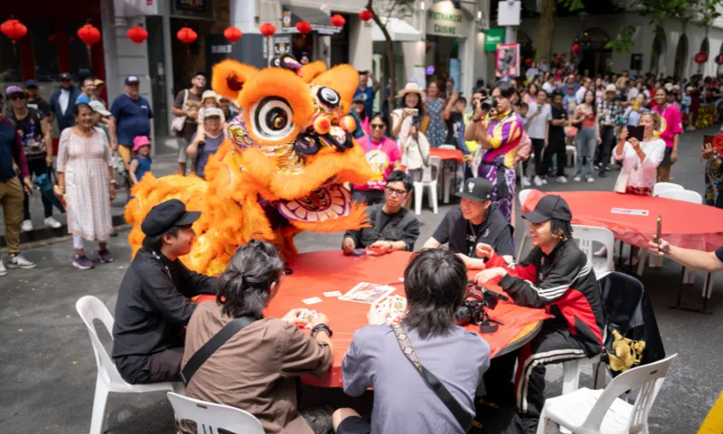 A photo of a table set up outside at a festival on a street. People are sitting around the table and looking at a large Chinese dragon (person in costume).