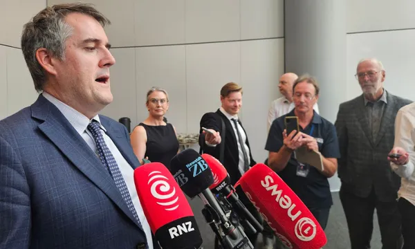 A man in a suit speaks at a press conference while several microphones and reporters with recording devices and notepads are positioned in front of him.