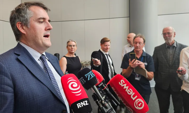 A man in a suit speaks at a press conference while several microphones and reporters with recording devices and notepads are positioned in front of him.