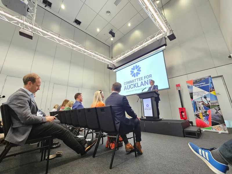 A speaker stands at a podium in a modern conference room, addressing an audience seated in rows. A large screen behind displays “Committee for Auckland.” People listen attentively, and banners are visible on the right.