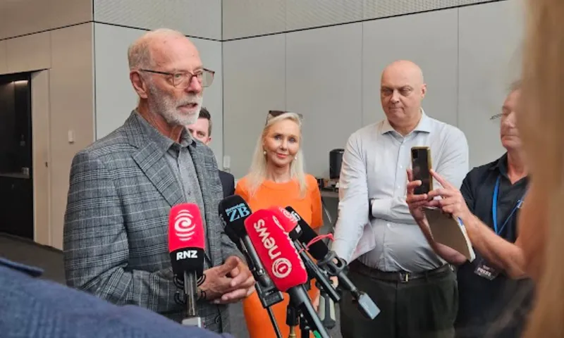 A man with gray hair and glasses speaks at a press conference, standing in front of several microphones. People, including a woman in an orange dress, listen and record with their phones in a modern indoor setting.