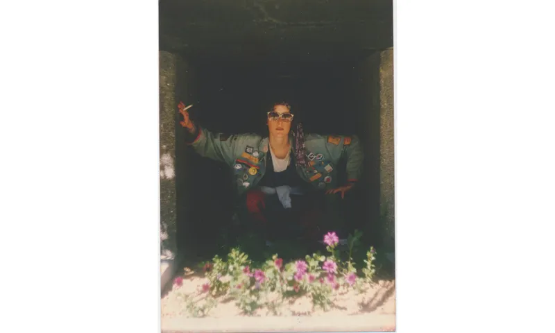 An old photo of a person wearing a denim jacket leaning out of a window, smoking, with an arrangement of flowers in front of them.