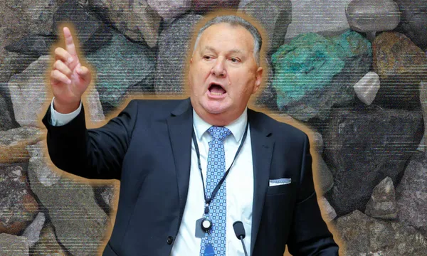 A man in a suit and tie stands in front of a background of large rocks, raising one hand and speaking with an open mouth.