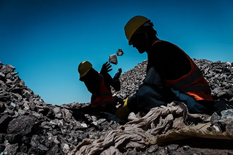 Two workers in hard hats and reflective vests are silhouetted against a blue sky, tossing rocks while sitting amid piles of stones at a worksite. Dust and debris are visible around them.