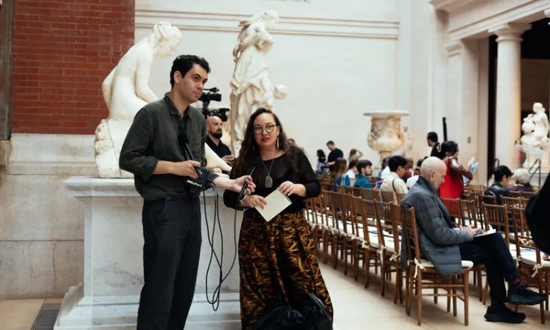 Two people stand holding equipment and papers in a museum-like setting with marble statues behind them. Several people are seated in rows of chairs, some using phones or talking, in the background.