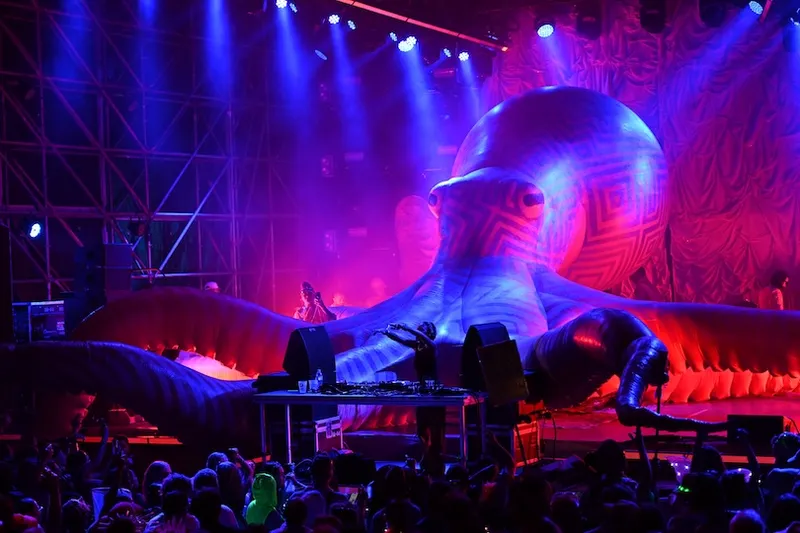A DJ performs on stage in front of a large, colorful inflatable octopus at a concert, with bright stage lights illuminating the crowd and creating a vibrant atmosphere.