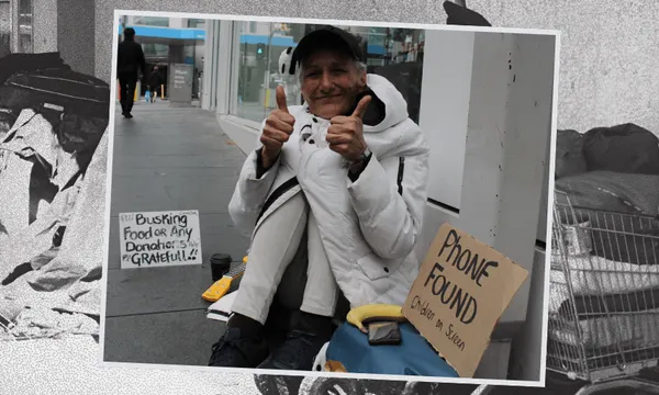 A smiling woman sitting on the sidewalk gives two thumbs up. Beside her are handwritten signs, one asking for donations for busking, and another stating "Phone found." Blankets and bags are visible around her.