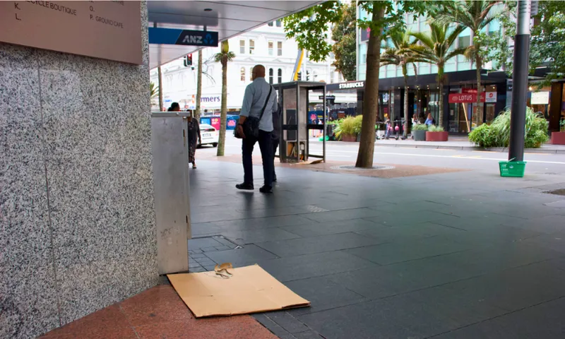 A man walks on a city sidewalk near a Starbucks, with a cardboard mat placed on the ground beside a building. Trees and shops line the street in the background.