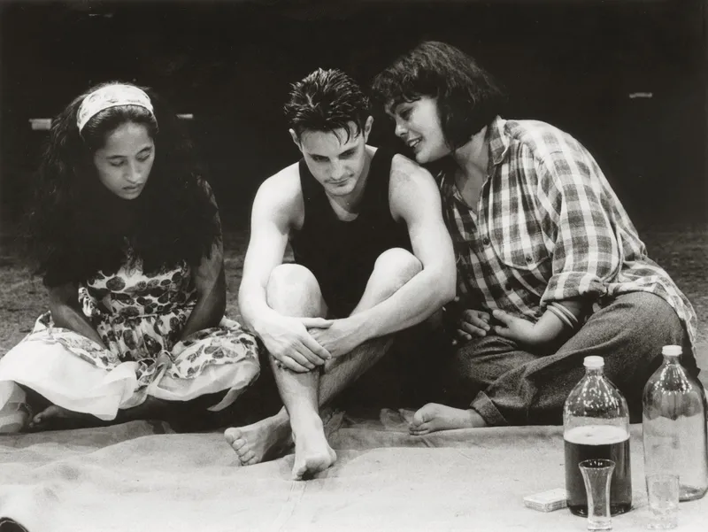 three young actors with dark hair sit on a stage during a play rehearsal. a man sits in a singlet between two women