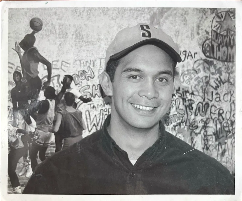 A young Māori man smiles at the camera wearing a dark collared shirt and a baseball cap