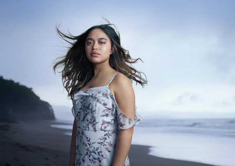 a young Māori woman with long dark hair stands on a gloomy beach wearing a light blue patterned dress