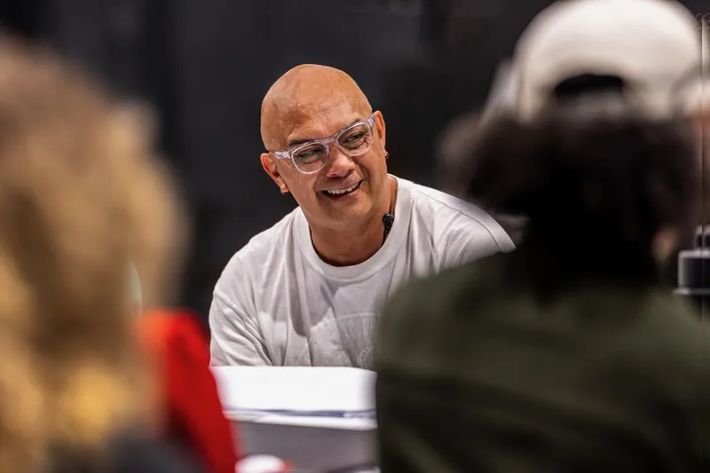 A middle-aged brown man with no hair and wearing glasses sits at a table, smiling in discussion with other people (out of frame)