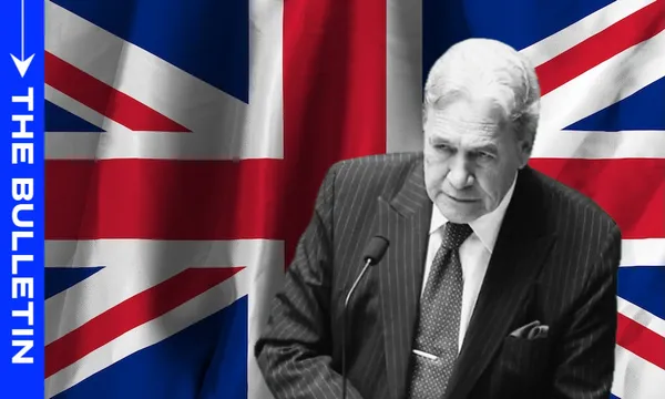 A man in a suit speaks at a podium in front of a large British flag; the left side has a blue vertical banner with the words "THE BULLETIN" in white text.