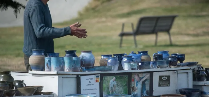 A photo of an outdoor table laden with ceramics. A person is standing behind the table, gesturing.