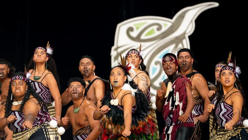 A group of Māori performers wearing traditional attire and facial markings perform a haka, expressing strong emotions, with a stylized design in the background.