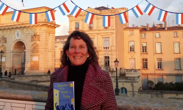 A photo of a woman standing in front of an historic building (French) in twilight. She is smiling and wearing a red coat. She is holding up a book.