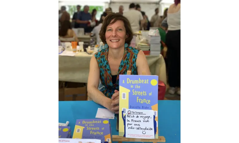 A photo of writer Daniella Blake who is sitting at a table in a marquee full of stalls. her book is propped up in front of her and she is smiling.