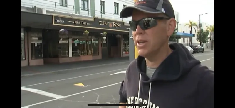 A man wearing sunglasses, a baseball cap, and a dark hoodie stands on a city street. In the background, there is a building with signs reading "Irish Pub" and "The Rose.
