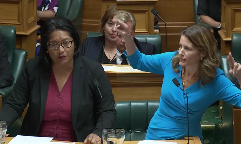 two female politicians debate in parliament, one is brown-skinned and wearing a red dress with a black blazer, the other is fair skinned and wearing a blue dress