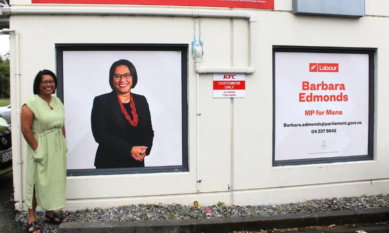 a woman in a green dress stands in front of a window sticker with her own portrait on it. to the right is another sticker reading "Barbara Edmonds, MP for Mana"
