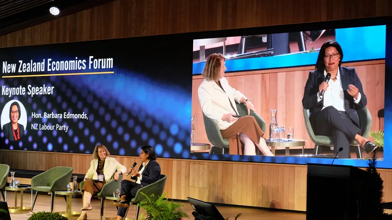 two women have a discussion on stage wearing business attire while a large screen behind them displays the name Barbara Edmonds and shows the two speaking