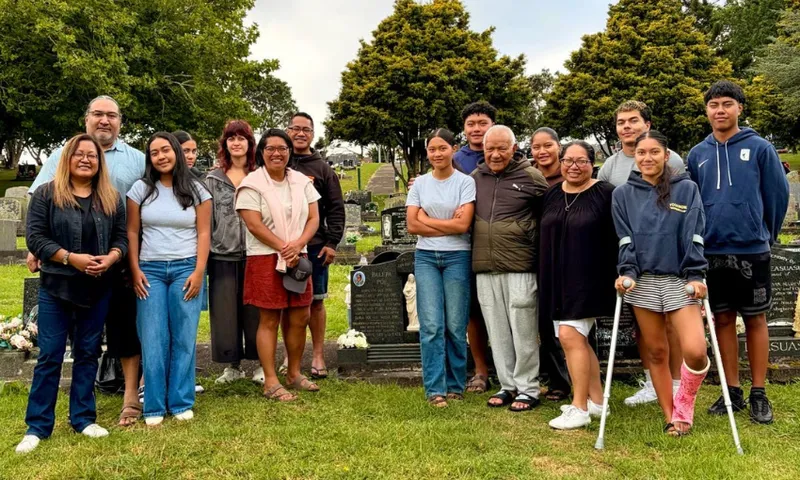 A large group of Samoan family members, young and old, gather around a black headstone at a cemetery and smile at the camera