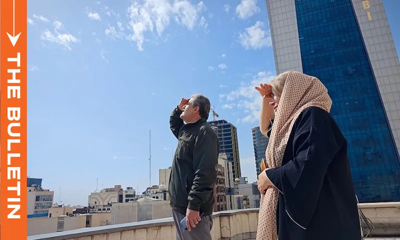 A man and a woman on a rooftop look up at the sky, shielding their eyes with their hands. Tall buildings and a blue sky with clouds are in the background. An orange column reads "THE BULLETIN" on the left side.