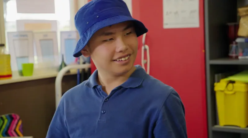A young person wearing a blue bucket hat and blue shirt smiles while looking to the side in a colorful classroom setting.