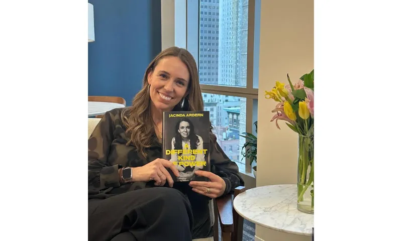 A photo of Jacinda Ardern sitting down and holding up a copy of her memoir. She is smiling and to her left is a low table with a bunch of flowers on it.