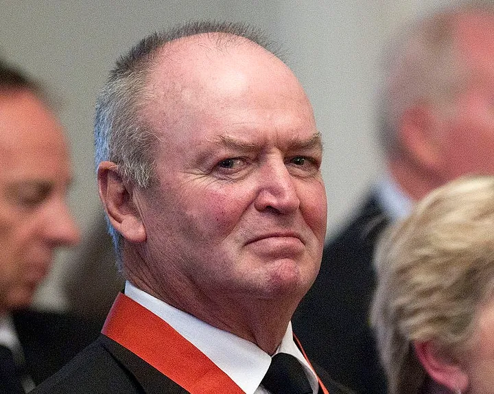 A man with short gray hair, wearing a black suit, white shirt, black tie, and red ribbon, stands in a formal setting among other people. He has a neutral expression on his face.