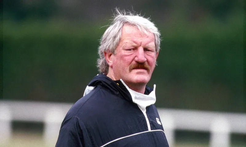An older man with gray hair and a mustache stands outdoors in a dark jacket with a half-zip collar. The background is blurred greenery.