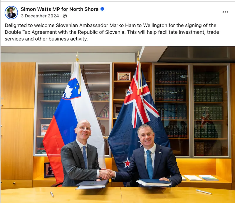 Two men in suits shake hands across a table, smiling, with documents in front of them. Behind them are the flags of Slovenia and New Zealand and shelves with books. The setting appears to be an official office or meeting room.