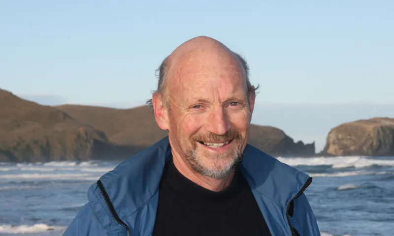 A photograph of Neville Peat who here is middle aged, standing on a beach wearing a jacket and smiling.