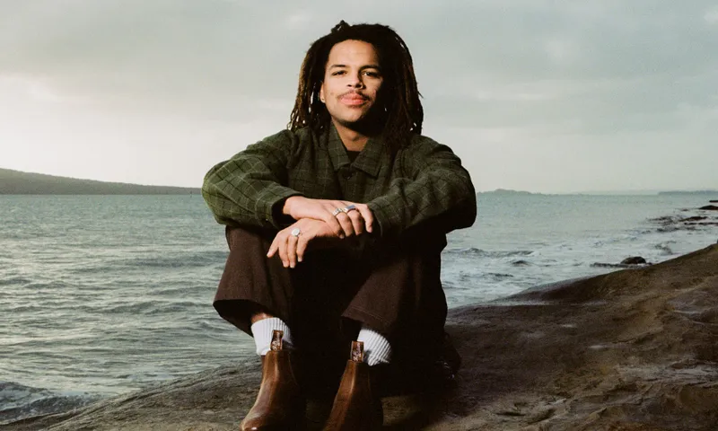 A photo of a man sitting by the ocean wearing warm clothes and hugging his knees. He has dreadlocks.