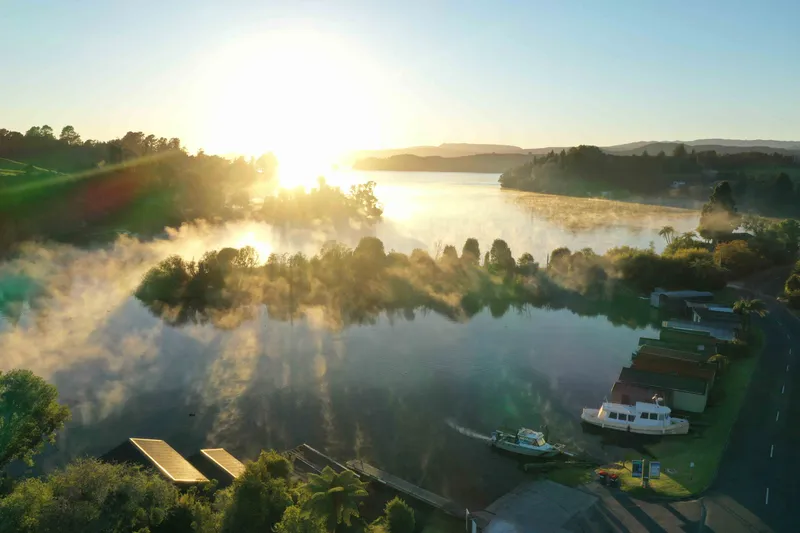 A serene lake at sunrise with mist rising from the water, surrounded by trees. Several boats and small docks line the shoreline on the right, and sunlight filters through the mist, creating a peaceful, golden atmosphere.